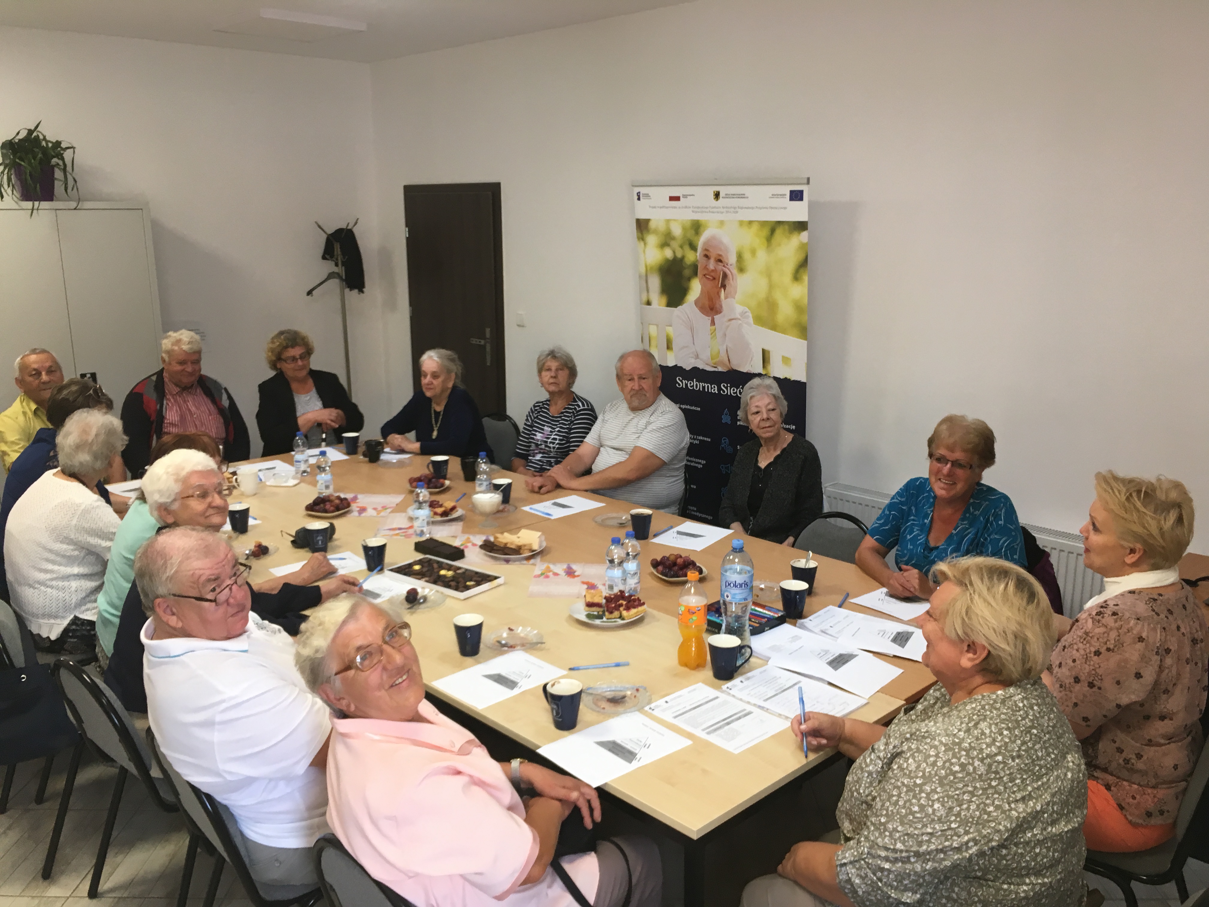 Seniorzy skupieniu wokół stołu w sali konferencyjnej ośrodka pomocy społecznej, wsłuchujący się w wypowiedzi prowadzących spotkanie trenerek.