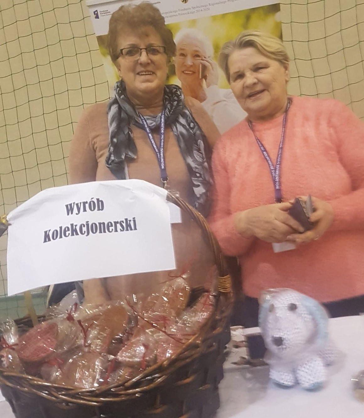 Wiklinowy kosz pełen pierniczków z załączoną kartką o treści: wyrób kolekcjonerski. W tle dwie panie promujące wypieki. 