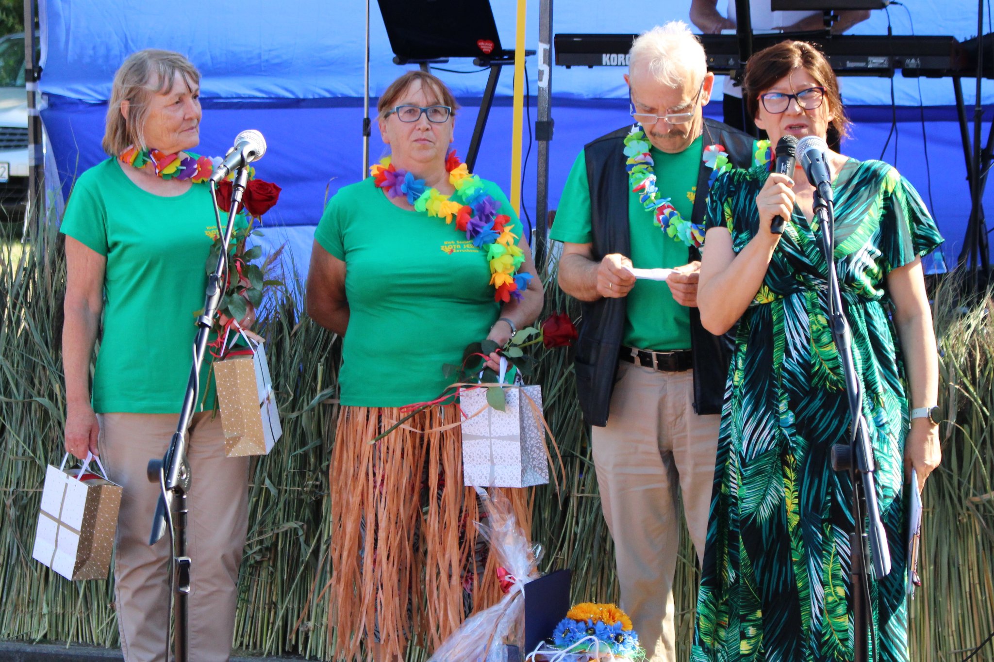Kierownik OPS przemawiająca do zgromadzonych, za nią trzech uczestników jednego z Klubów Seniora z upominkami  